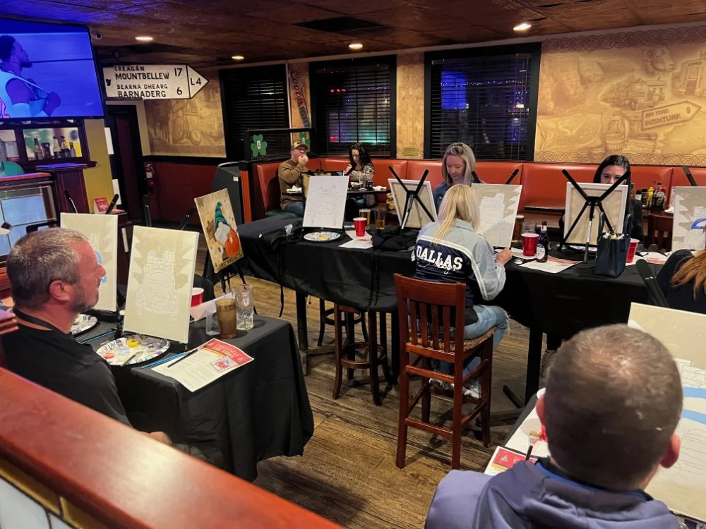People participating in a group painting class in a restaurant, sitting at tables with painted canvases in front of them.