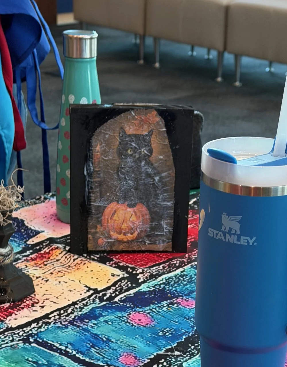 A small sign with an image of a black cat sitting inside a carved pumpkin, with a moon and stars background, on a colorful tablecloth. There is a blue water bottle and a Stanley tumbler on the table, along with other items.