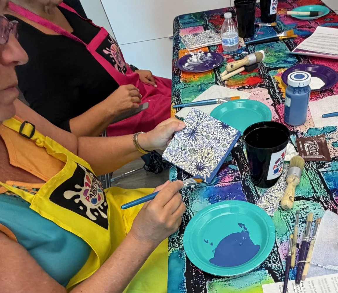 People sitting around a table painting tiles at a paint and sip event, with various paints, brushes, and supplies on the colorful table.