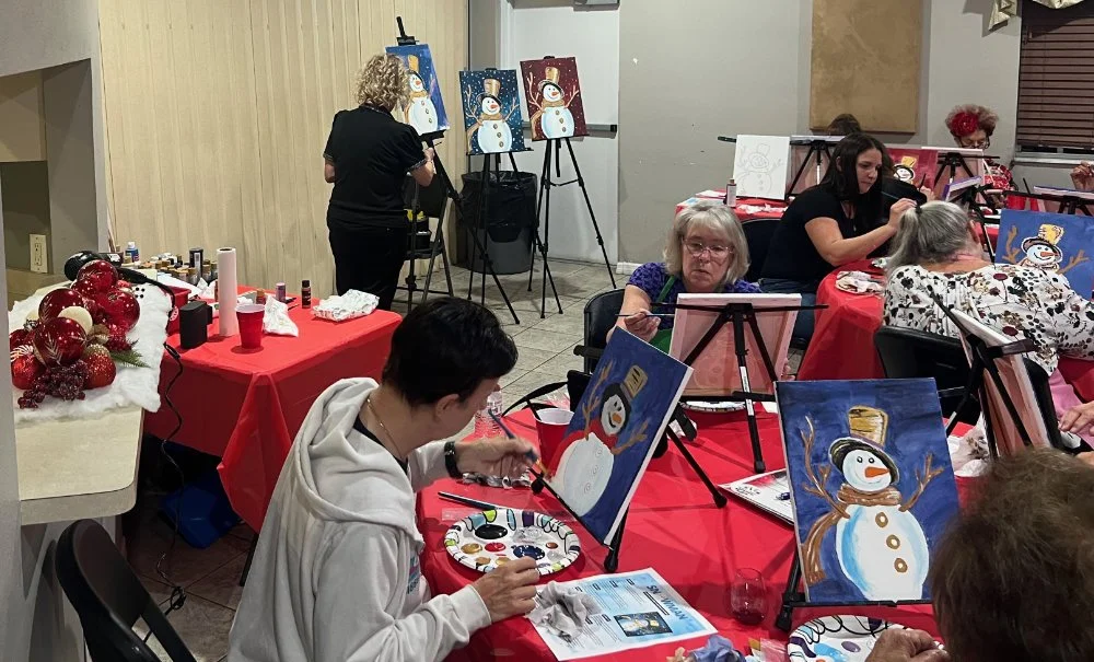 Group of people painting Christmas snowman pictures at an indoor art class with red tablecloths and holiday decorations.