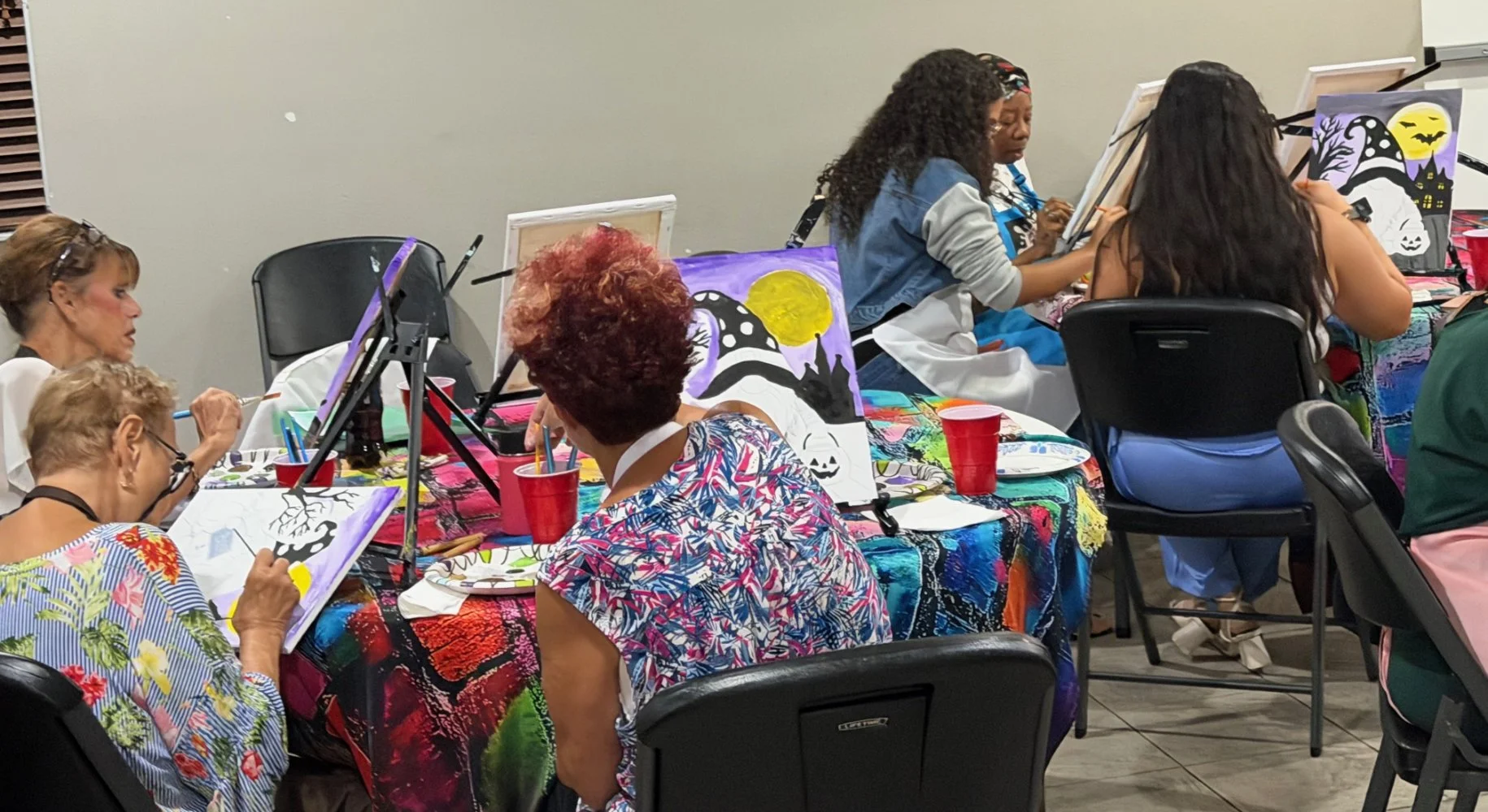 Several women participate in a Halloween-themed painting event, creating paintings of a spooky scene with a haunted house, bats, and a full moon.