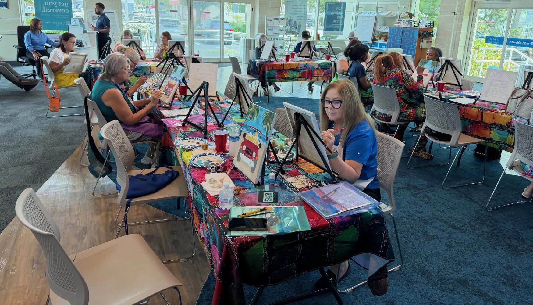 People participating in a painting class at a colorful studio, seated at tables with canvases and art supplies, with a large glass window in the background showing outside signage and parking lot.