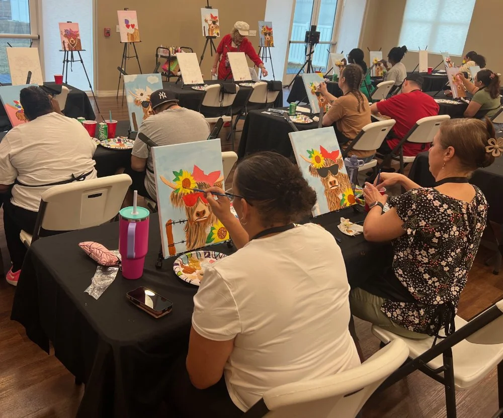 People participating in a painting class, creating artwork of a llama wearing sunglasses and a floral headpiece, in a well-lit room with large windows and various finished paintings displayed on easels.
