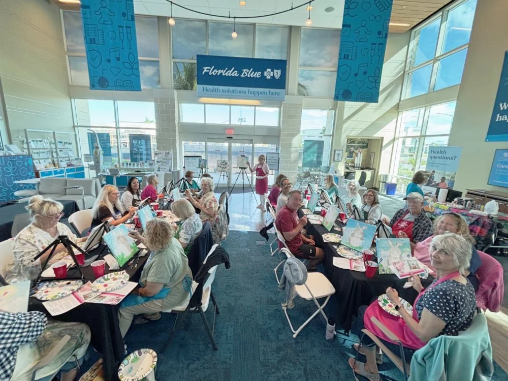 A group of people participating in a painting class at Florida Blue, seated at long tables with painting supplies, in a bright, spacious room with large windows and blue-themed decorations.
