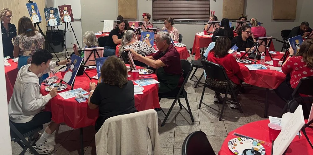 People of various ages gathered around tables with red tablecloths, painting snowmen on canvases during a group art activity in a well-lit room.