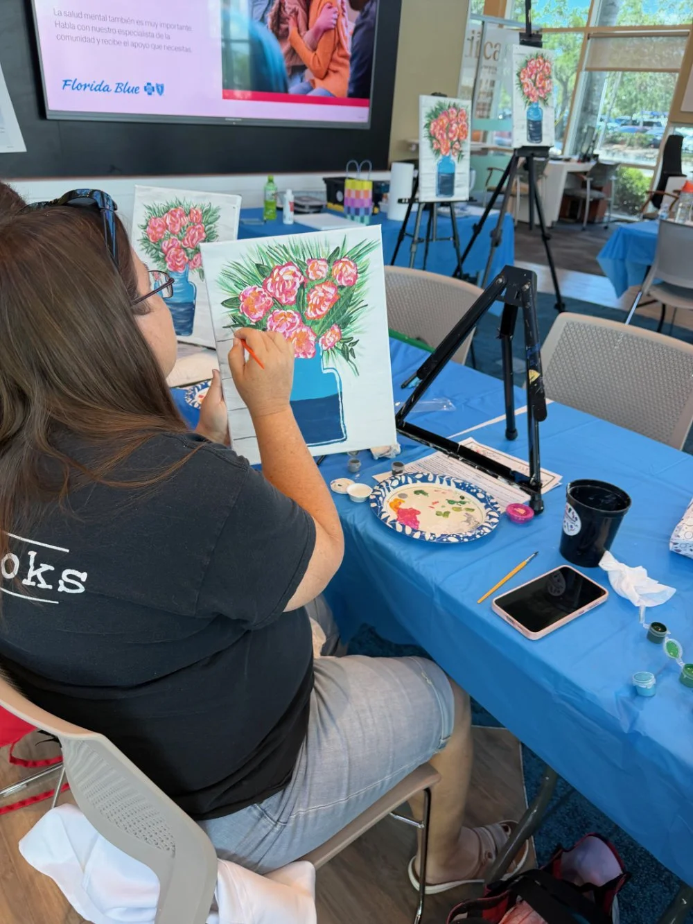 A woman painting a colorful floral arrangement on canvas at a painting class, seated at a table with painting supplies and snacks, in a room with large windows and art displayed on easels.