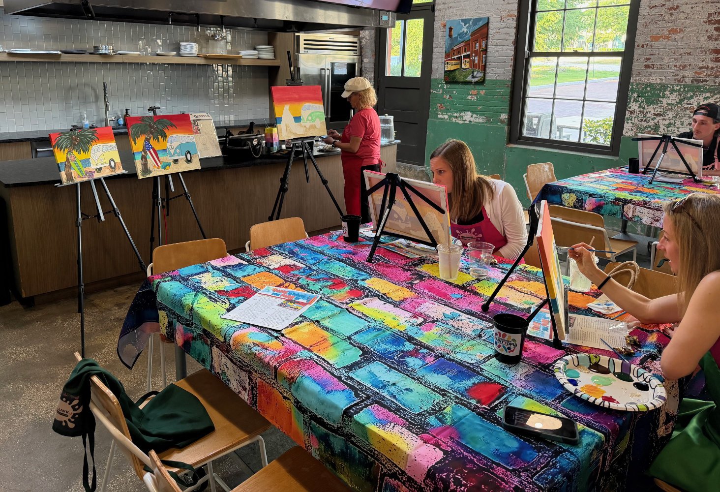 A group of women participating in a painting class inside a rustic, colorful art studio, with some paintings on easels and completed art on the tables.