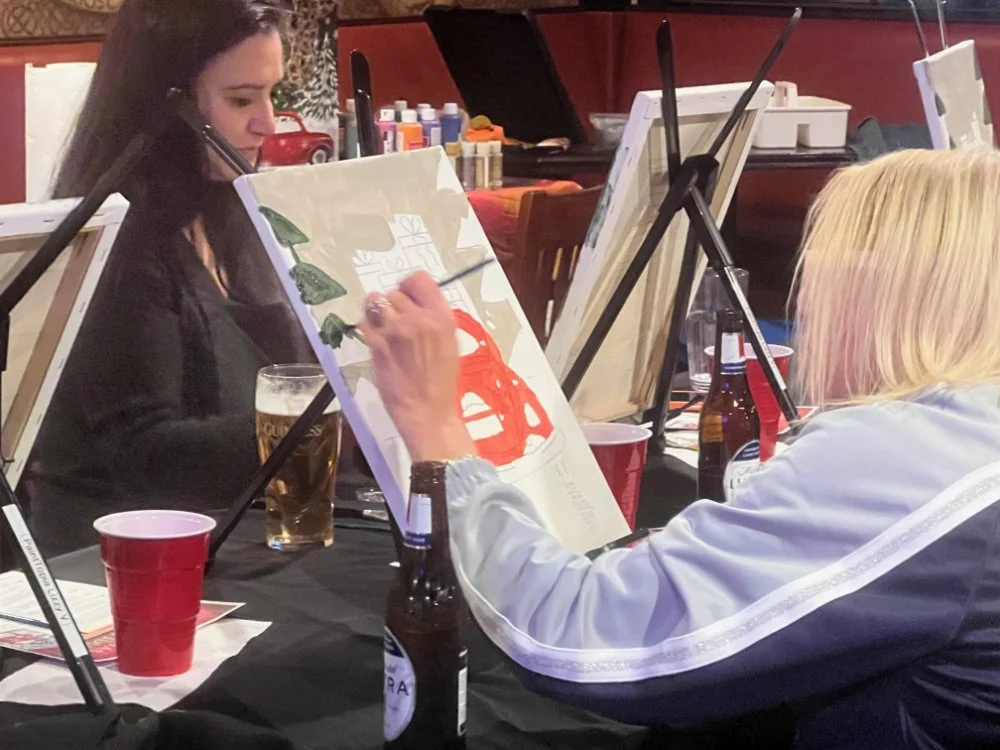 Two women painting on canvases set on easels in a crowded indoor setting, with bottles and red cups on the table.