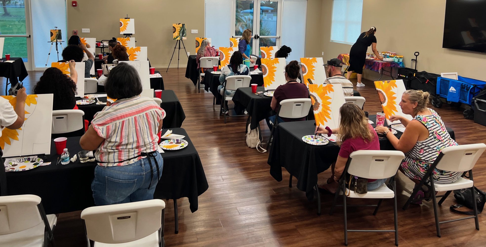Group of people in a art class, painting sunflower pictures on canvases at black-covered tables, with a window in the background and a woman arranging supplies at a table to the right.