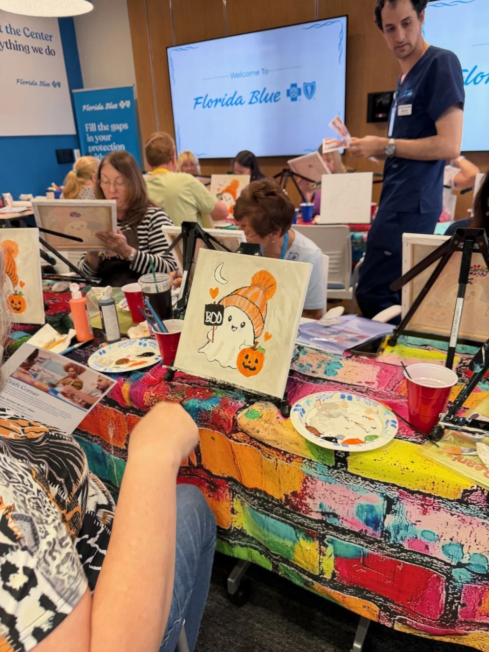 Group of people participating in a painting activity at a colorful table with Halloween-themed artwork, including a ghost with a pumpkin hat. A large screen in the background displays 'Welcome to Florida Blue' in a classroom setting.