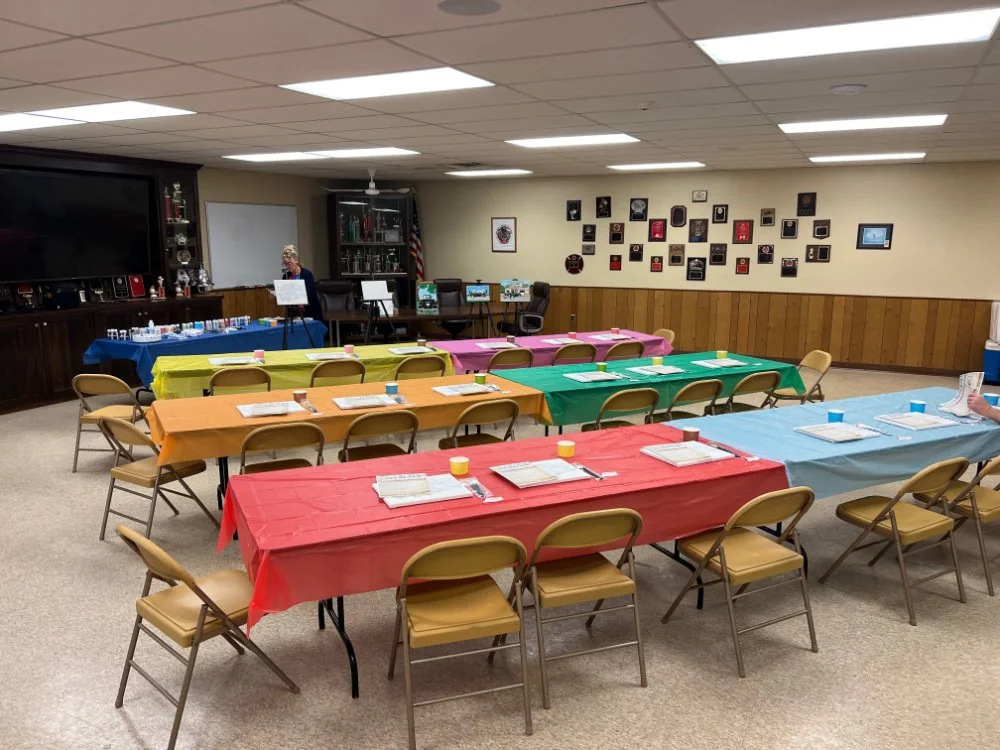 A decorated room set up for a gathering with tables covered in colorful tablecloths, each with plates, napkins, and cups arranged for guests. There is a display table at the back with awards and trophies, and a person standing near it. The walls have