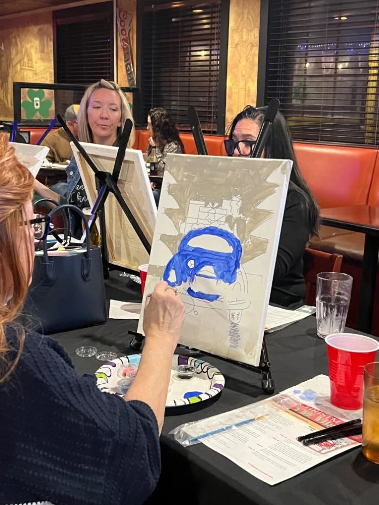 People painting a portrait on a canvas in a restaurant. The canvas shows an outline of a face wearing glasses, being painted in blue.
