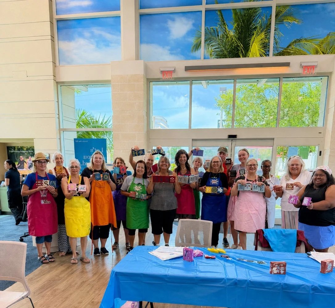 Group of women wearing colorful aprons standing inside a bright indoor space with large windows, holding decorated blocks that spell out 'Home Sweet Home'.