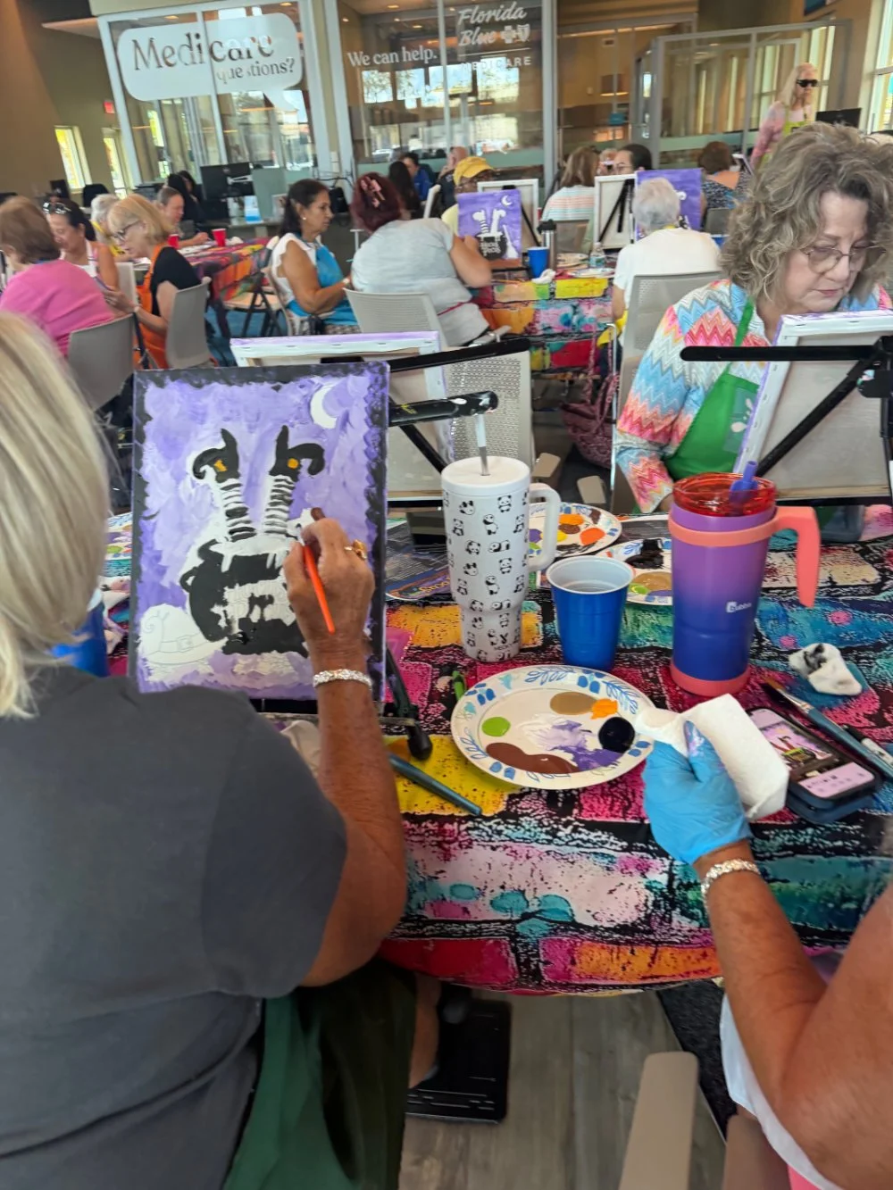 A woman painting a unicorn on a canvas during a group art activity inside a lively, colorful event space with many people engaged in similar activities.