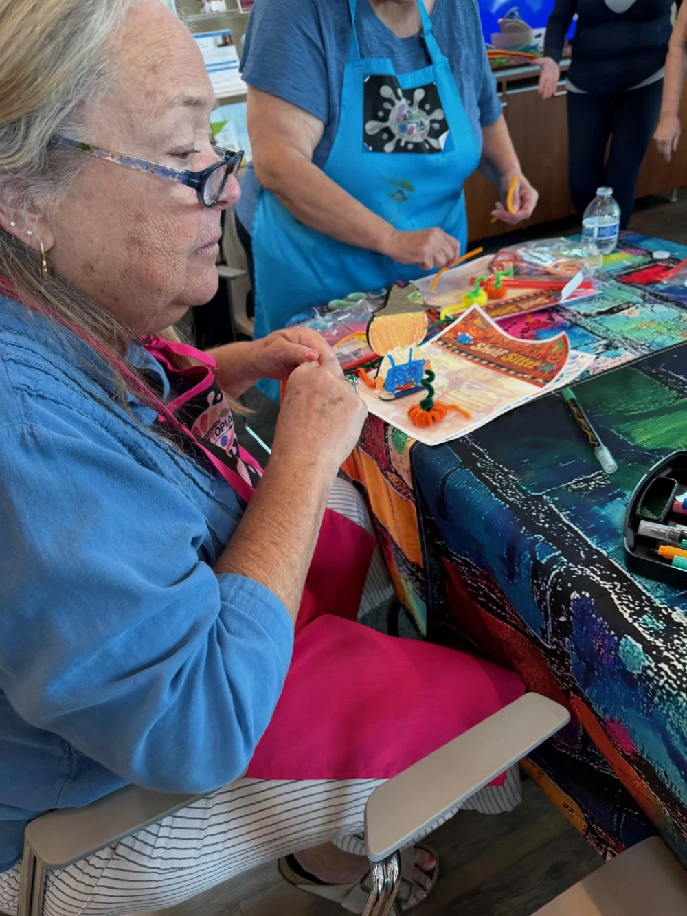 An elderly woman with glasses and a pink apron sitting at a table, creating a craft project with colorful pipe cleaners and craft materials, with another person in a blue apron standing nearby, in a setting that appears to be a craft or activity even