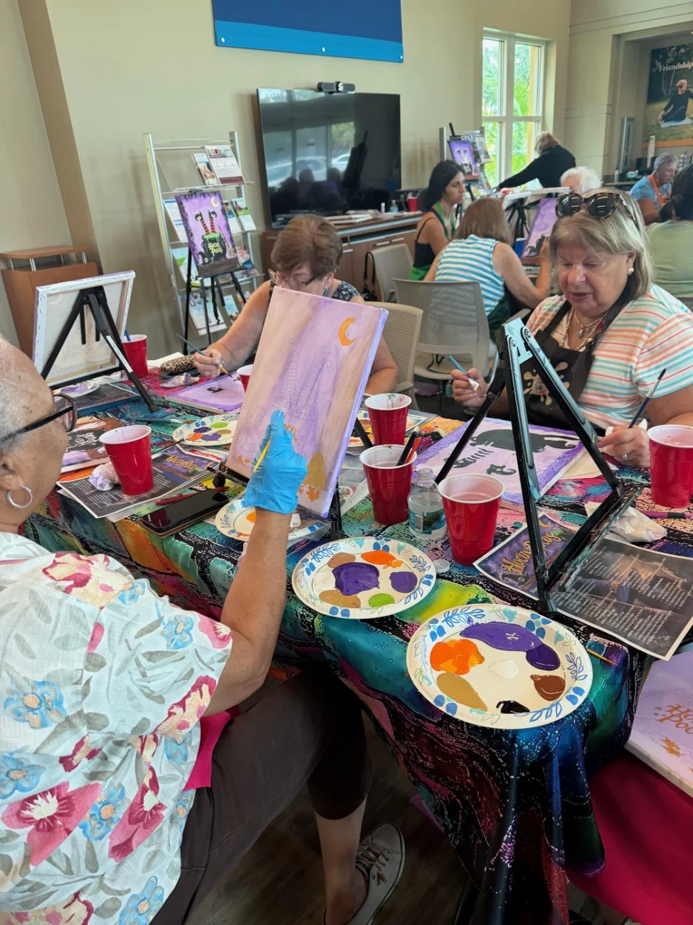 Group of women participating in a painting class indoors, sitting at a table with painting supplies, easels, and canvases, with some paintings depicting night scenes with moons and animals.