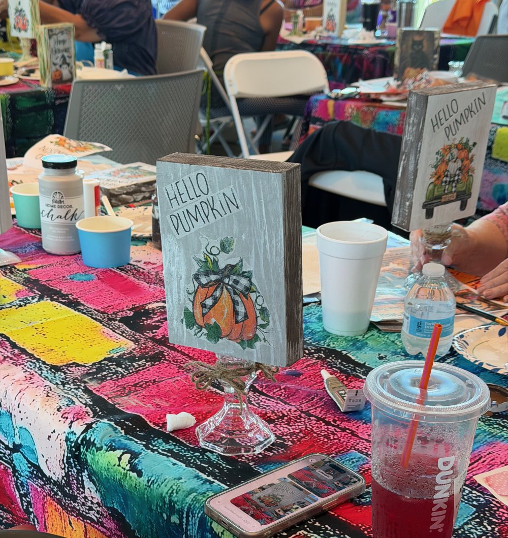 Table decorated with colorful tablecloth at a craft or party event, featuring signs with pumpkins and autumn-themed decorations, disposable cups, a smartphone, and various craft supplies.