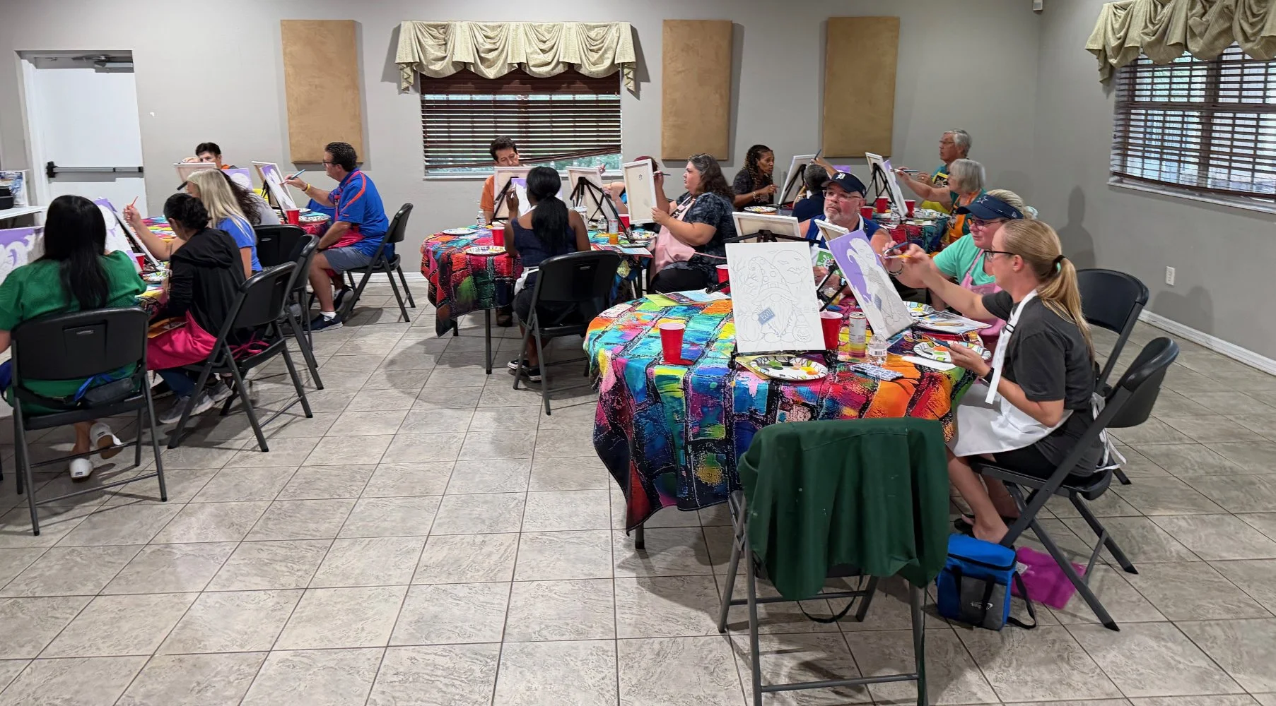 A group of people participating in a painting class, seated at tables covered with colorful tablecloths, each with a canvas, brushes, and paints, in a room with windows and beige curtains.