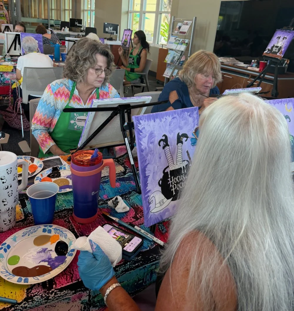 A group of women engaged in a painting class, painting a Halloween-themed picture of a witch flying on a broomstick with a purple background and crescent moon. The scene features art supplies, easels, and a large television in a well-lit room.