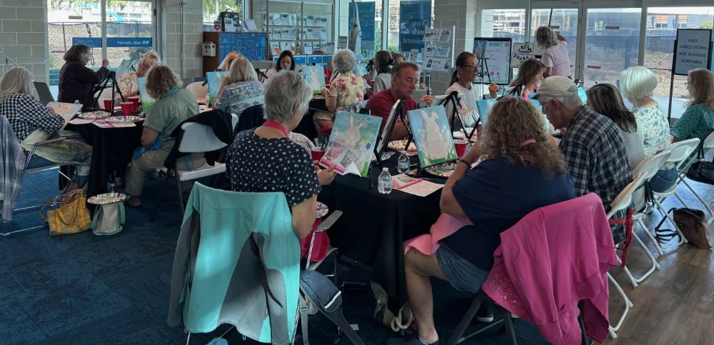 People participating in a group painting class or art workshop in a bright, airy room with large windows.