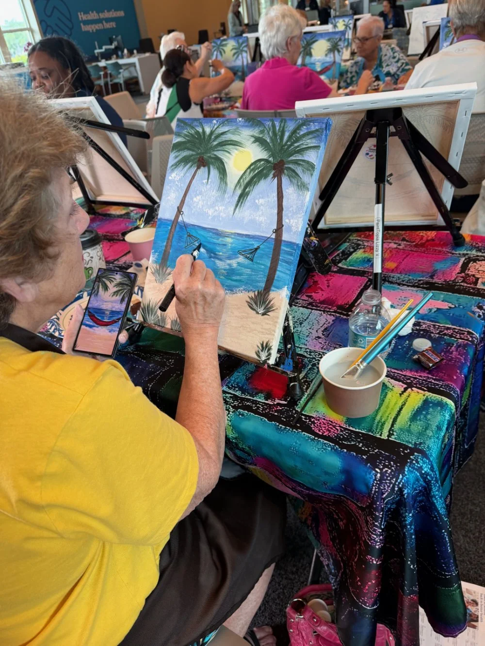 An elderly woman painting a tropical beach scene with palm trees, swings, and a sun on a canvas at a group painting event.