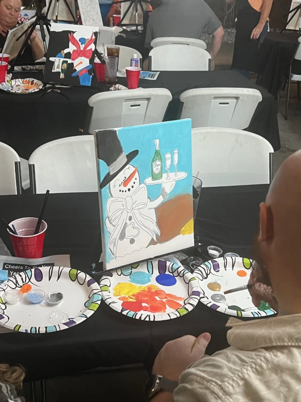 A person with glasses and a beige shirt is looking at a painting of a snowman holding a tray with a bottle and glasses. The painting is on a table with plates of paint, cups, and brushes, at an event with other people in the background.