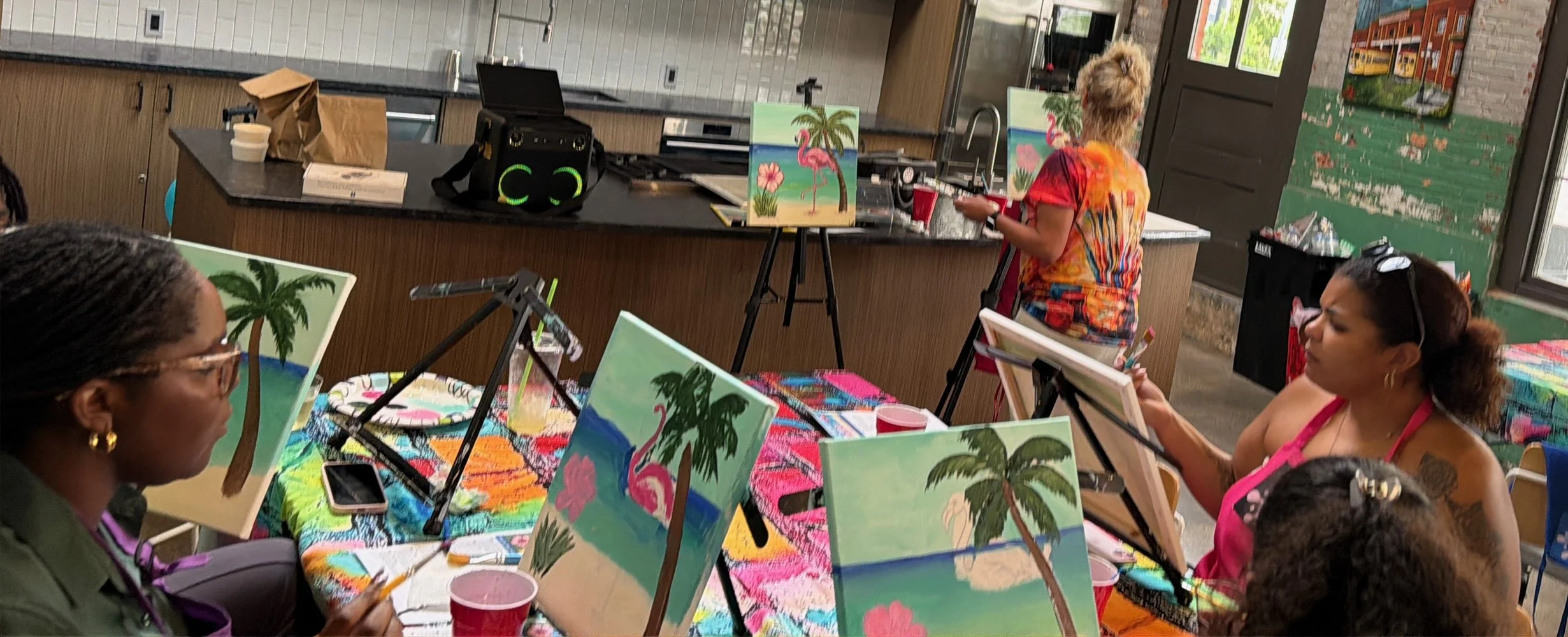A group of people participating in a painting class with tropical beach scenes, featuring palm trees and pink flamingos, set up on easels around a table in a room with large windows and colorful decor.