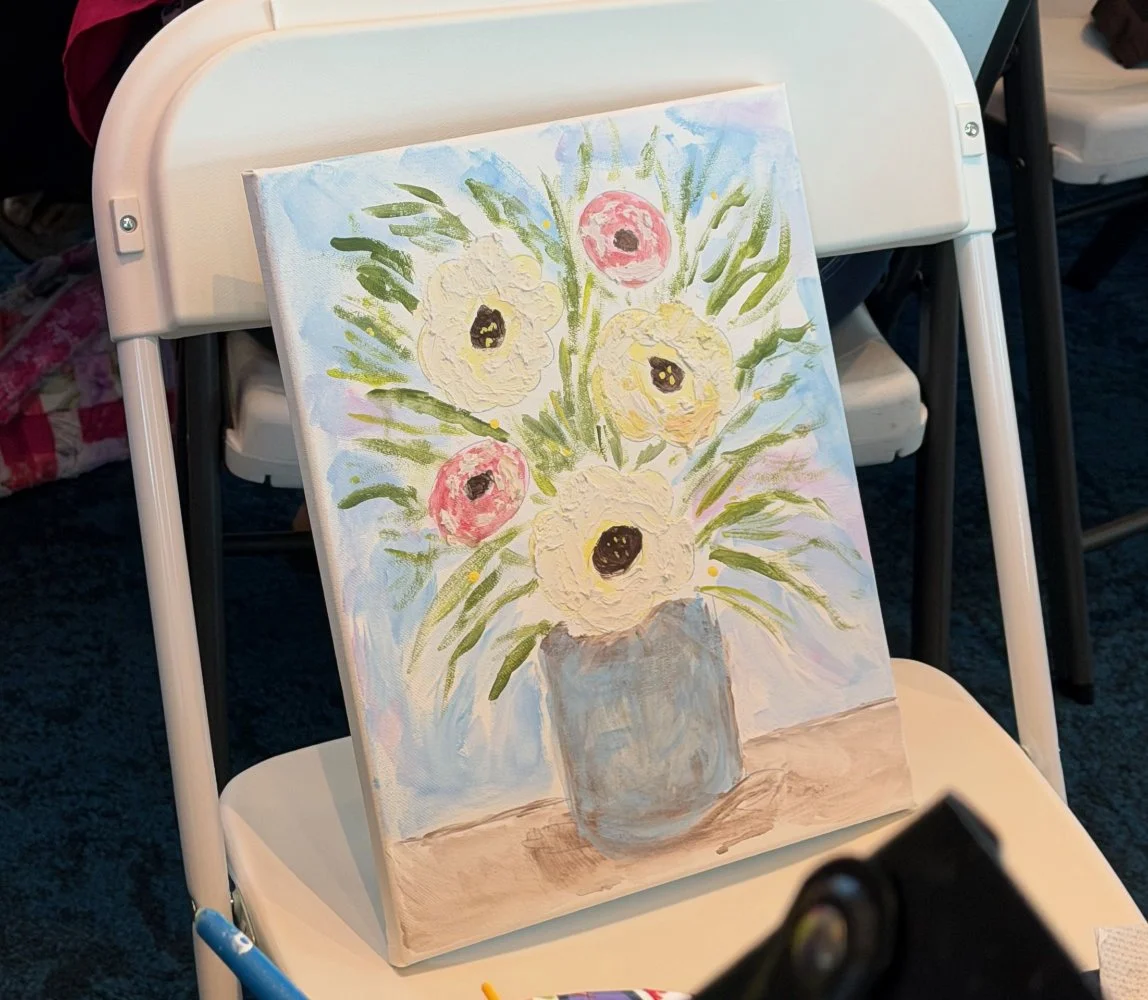 A painting of a bouquet of flowers in a vase, placed on a table, resting on a white chair.