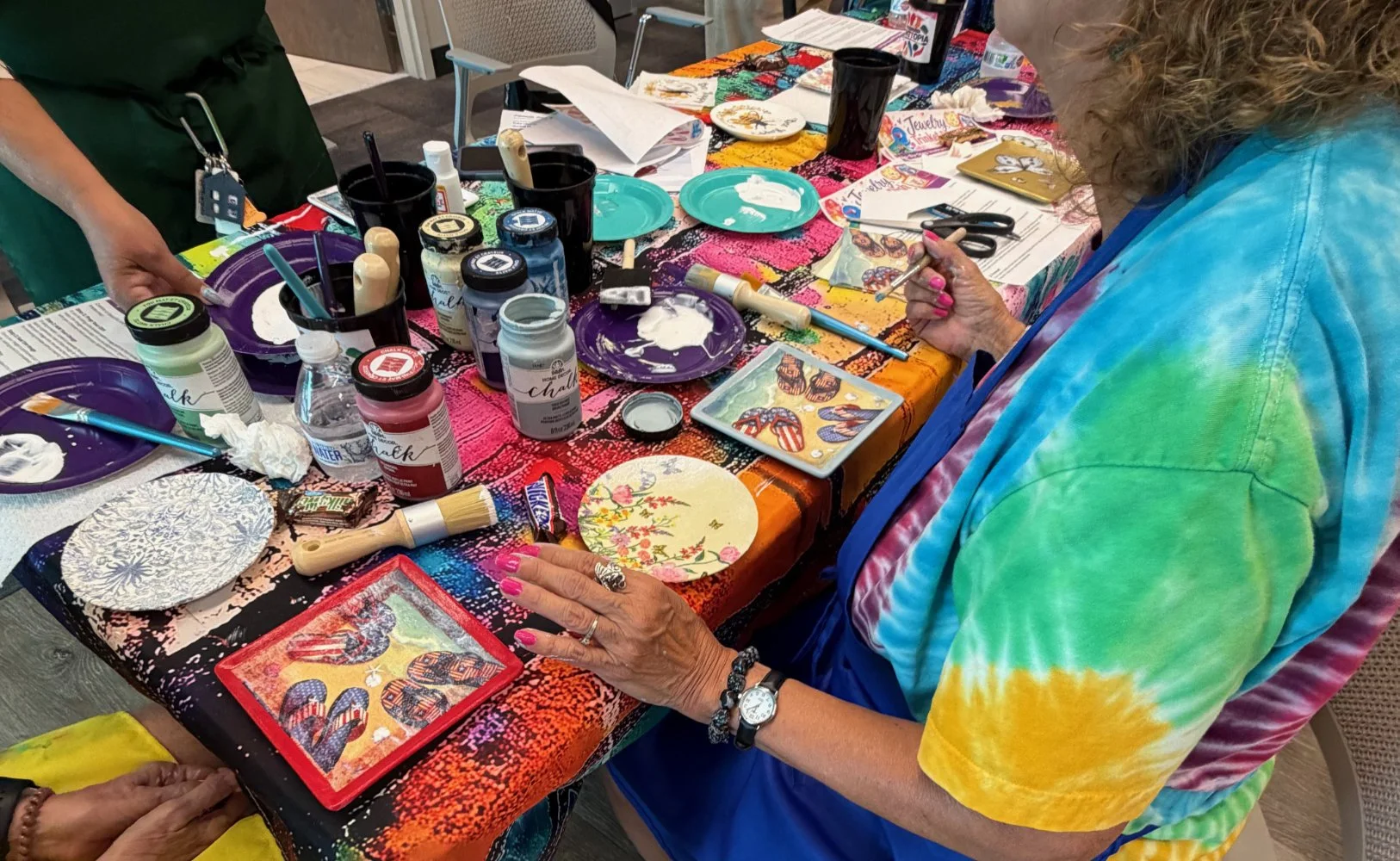 A woman in a colorful tie-dye shirt is painting on a small square canvas at a craft table, surrounded by paints, brushes, paper plates, and other art supplies.