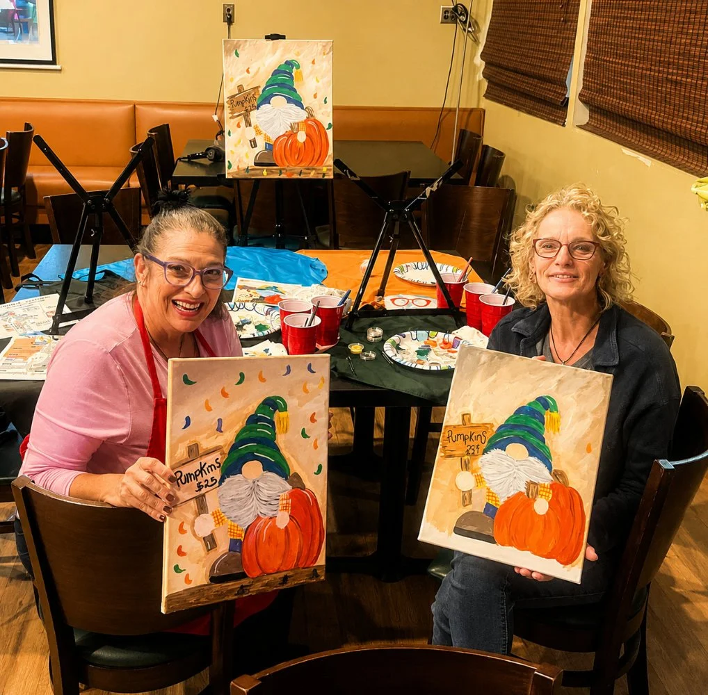 Two women smiling and holding painted canvases of a gnome with a pumpkin, in an art studio. The table has painting supplies and red cups, with additional completed paintings on a table behind them.