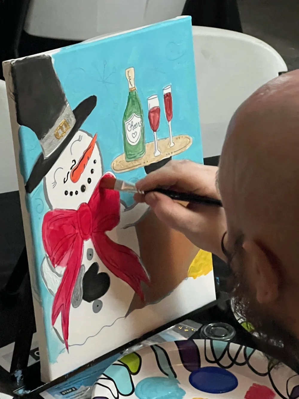 Person painting a festive snowman with a black top hat, red bow, and carrot nose, on a colorful canvas featuring a bottle of champagne and two glasses of red drink.