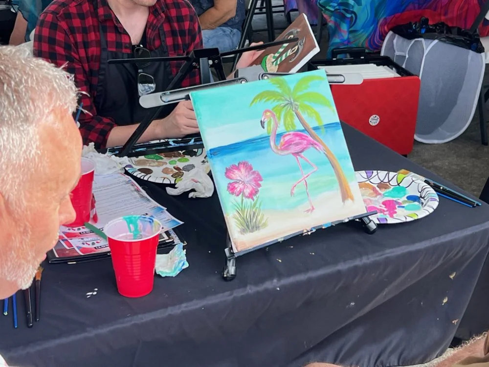 A painting of a pink flamingo standing next to a palm tree under a blue sky on a beach scene.