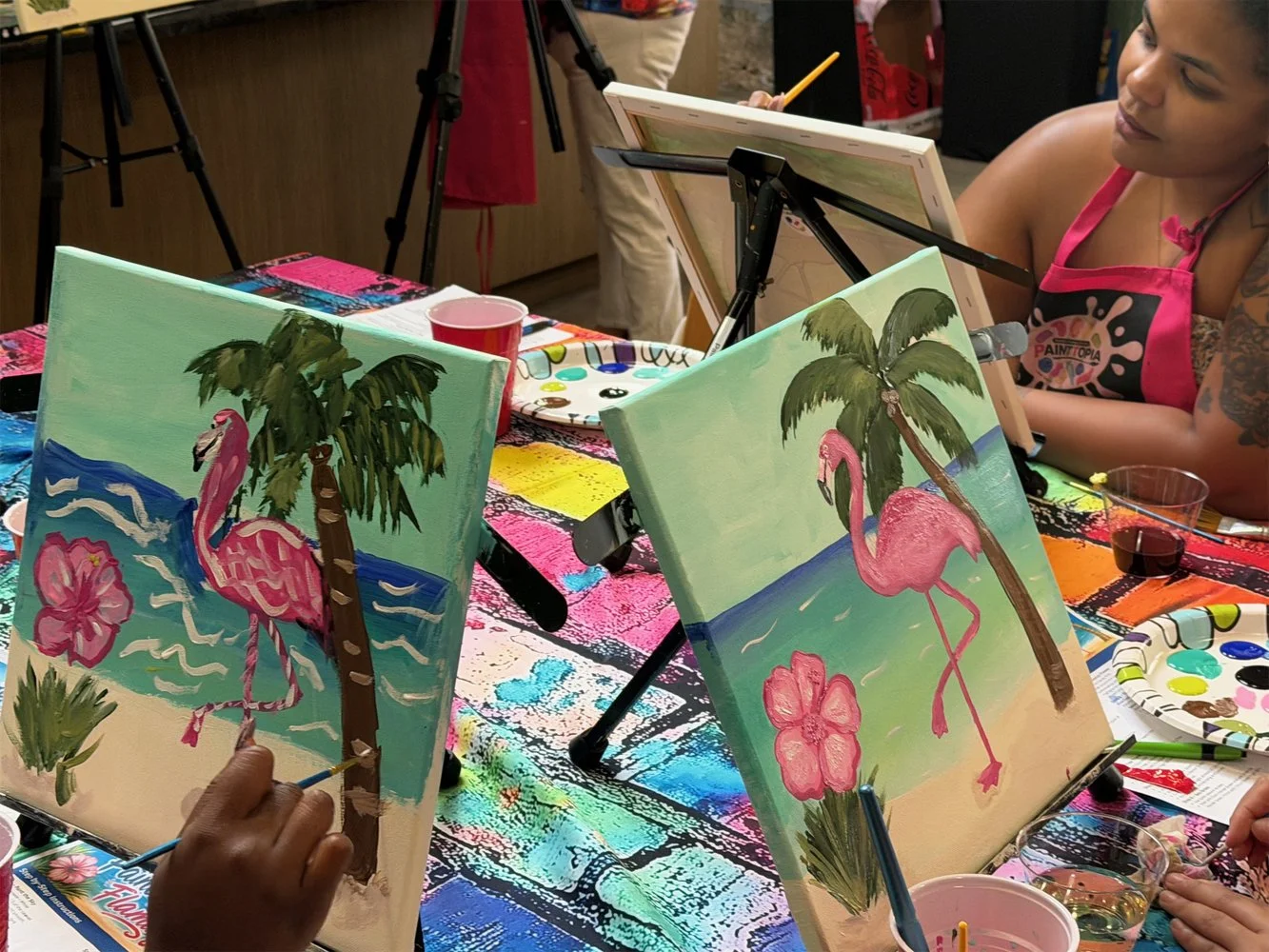 People painting tropical scene with pink flamingos, palm trees, blue water, and flowers on canvas at an art class.