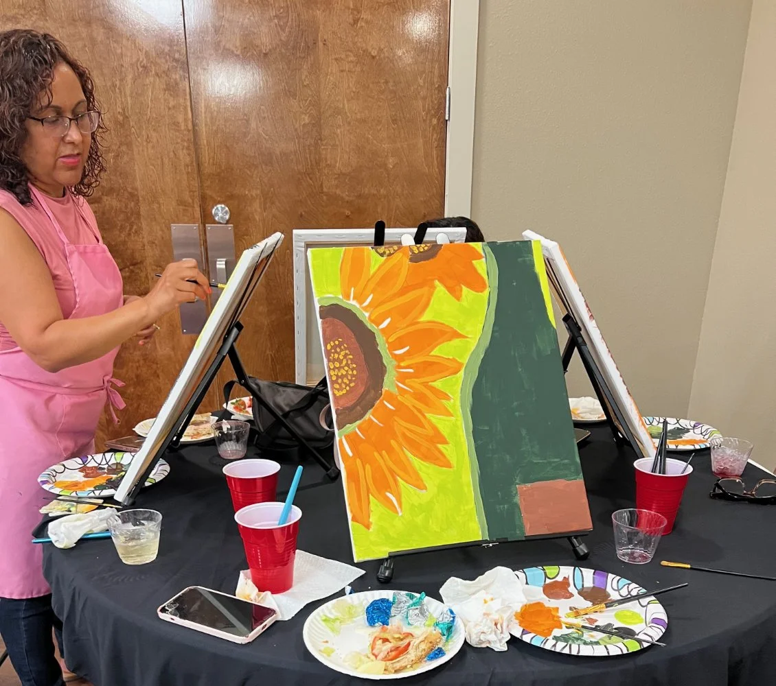 A woman wearing glasses and a pink apron paints a sunflower on a canvas at a paint-and-sip event. The table is scattered with paint supplies, cups, and paper plates with paint.