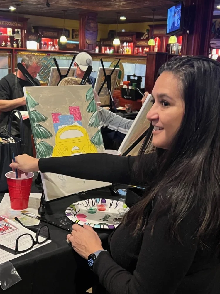Woman with long dark hair painting a holiday-themed canvas in a restaurant with others painting at tables, Christmas decorations, and a TV screen in the background.