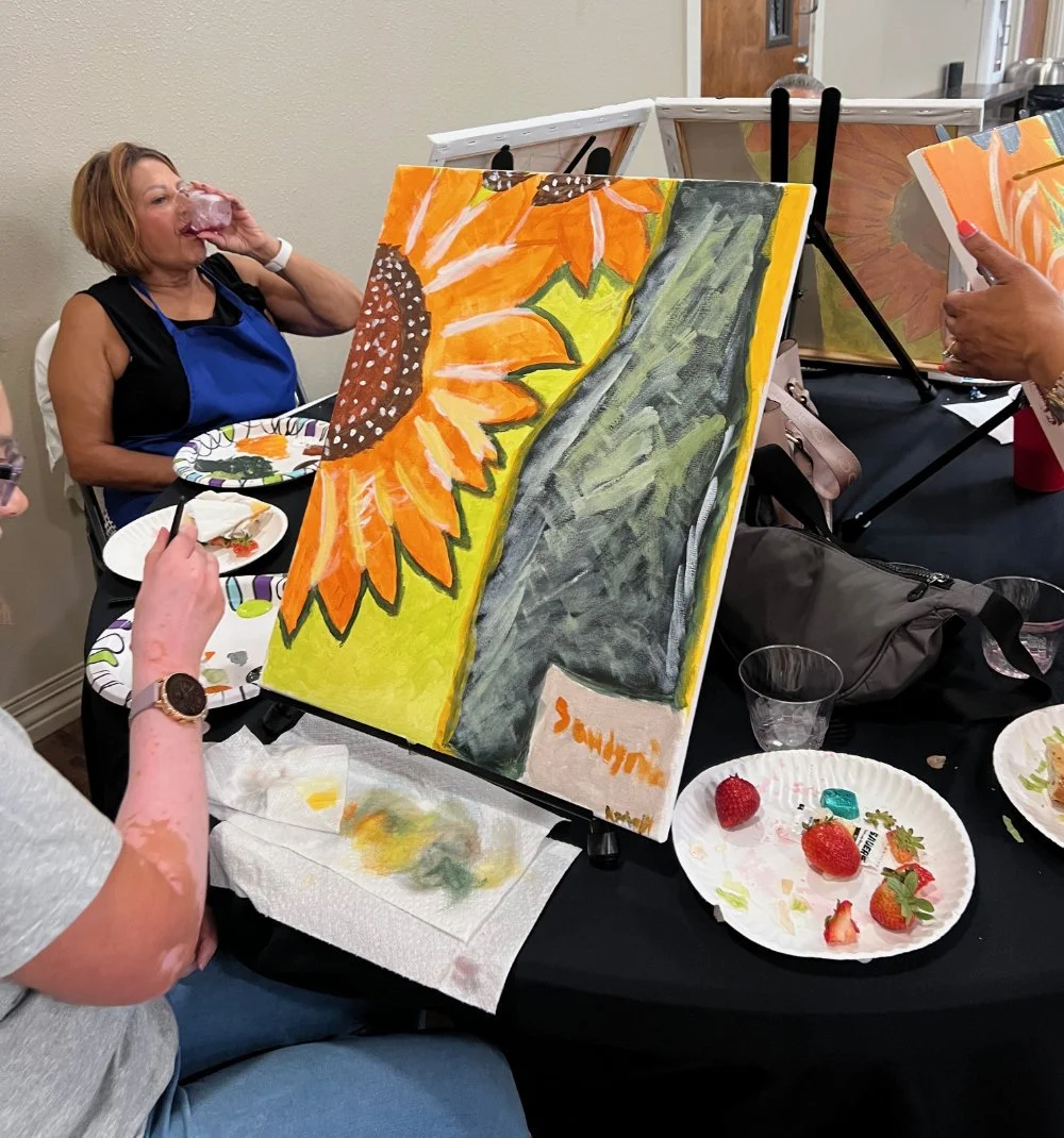 A woman at a paint and sip event is painting a sunflower on a canvas. She is drinking from a glass and wearing a blue apron. There are other people, art supplies, and strawberries on plates on the table.