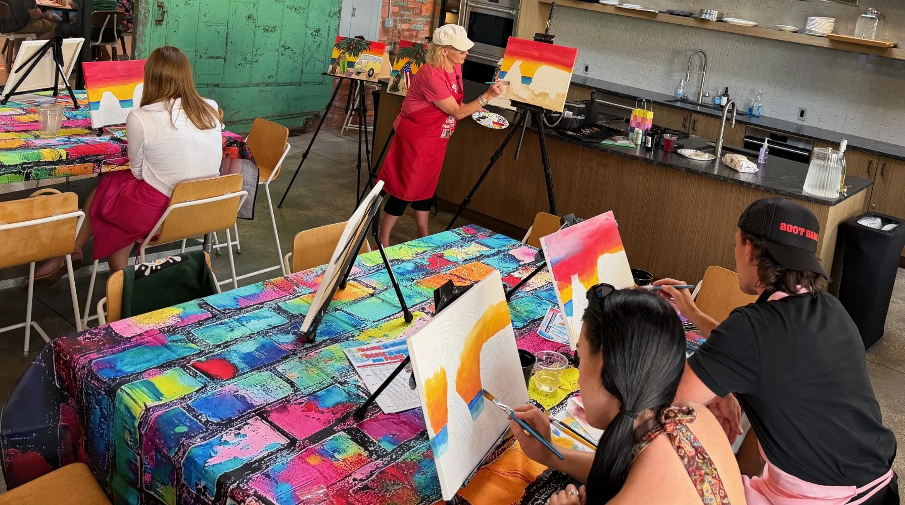 Group of people in a colorful painting class, painting sunset scenes on canvases, with a woman in a red apron guiding them.
