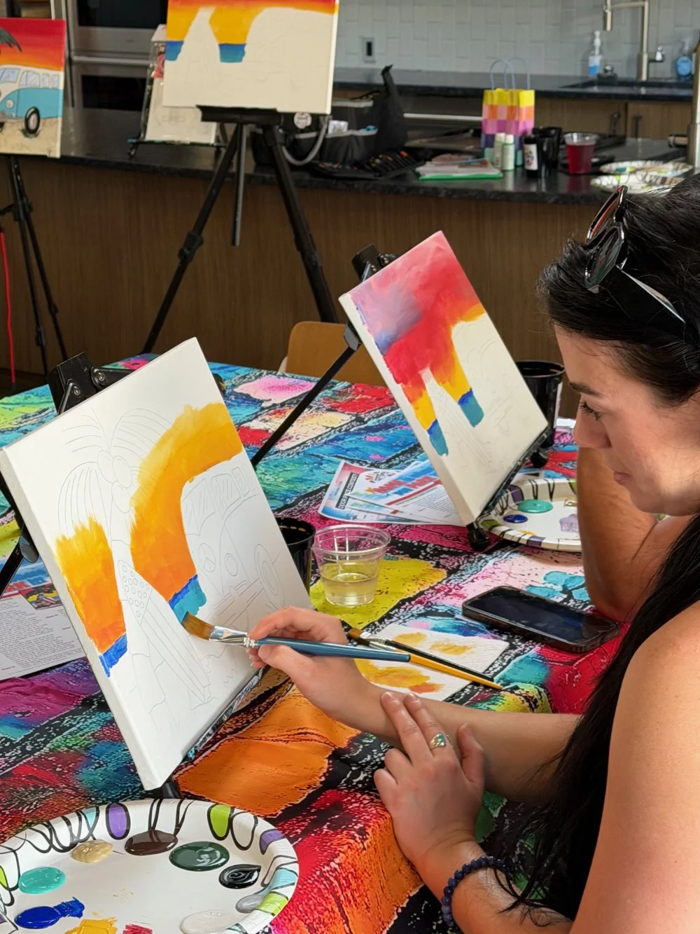 A woman is painting a colorful abstract art piece on a canvas at a paint and sip event. There are multiple canvases on easels, paint supplies, and a vibrant tablecloth in the background.