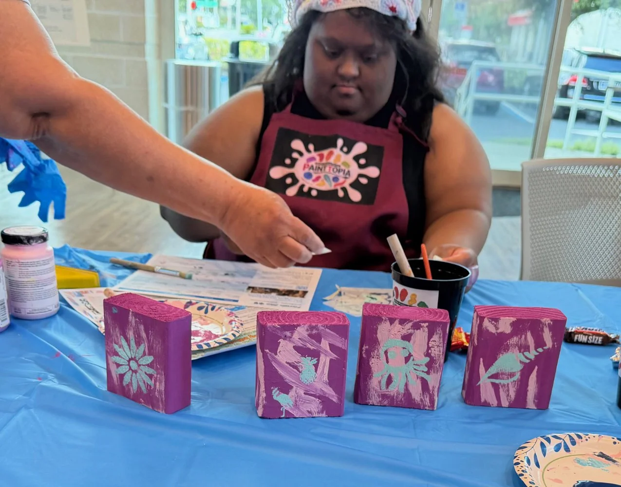 A young girl wearing a paint apron and headband decorated with red stars is painting on four wooden blocks. An adult's hand is helping with her art project. The table is covered with blue plastic and is set with painting supplies, including paint bot