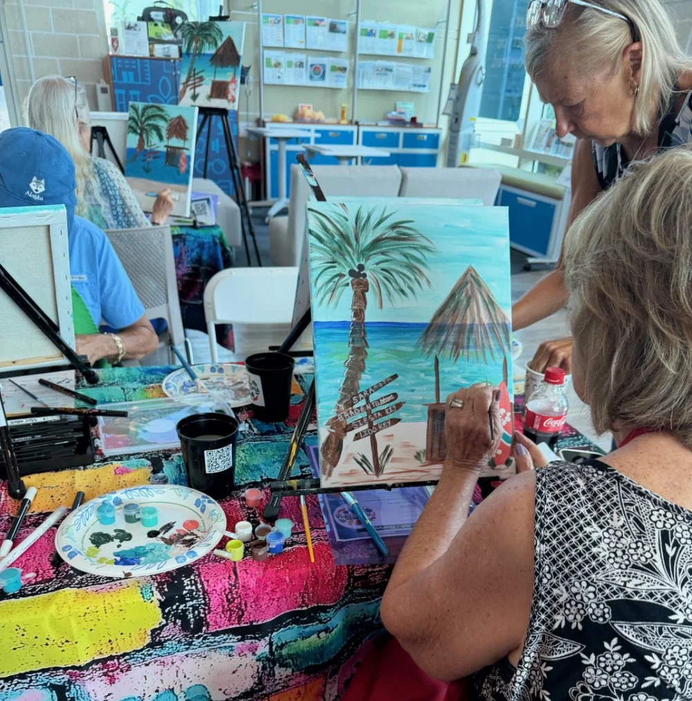 Older women painting a tropical beach scene with palm trees and umbrellas at a paint and sip class.