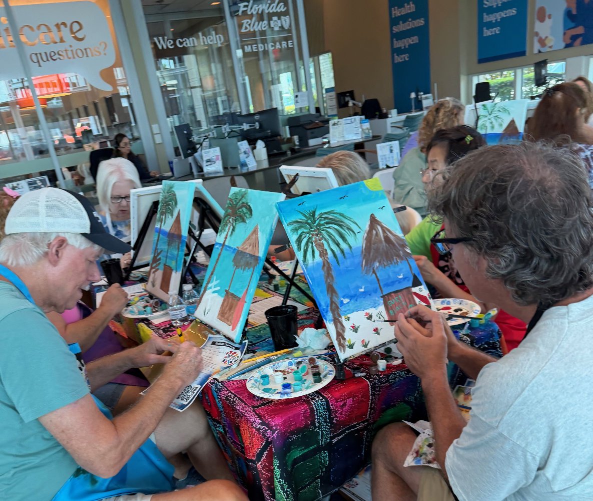 People participating in a group painting activity at a table with colorful paintings of tropical beach scenes with palm trees and mountains, inside a lobby area with large windows and signs promoting healthcare and Medicare services.