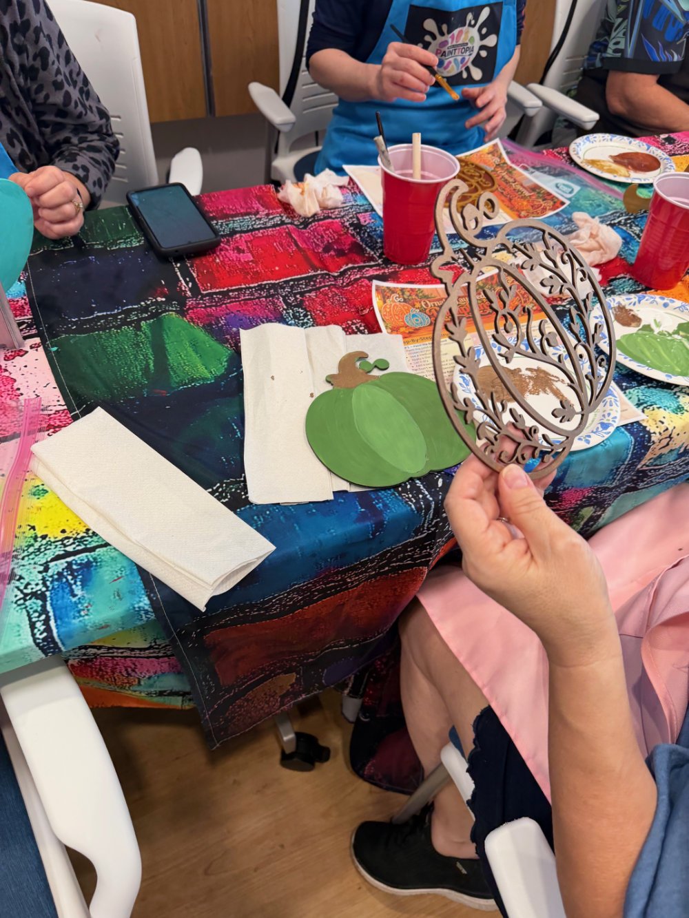 People sitting around a table with colorful tablecloth, craft supplies, paper pumpkins, and paper cutouts, engaging in a craft activity.