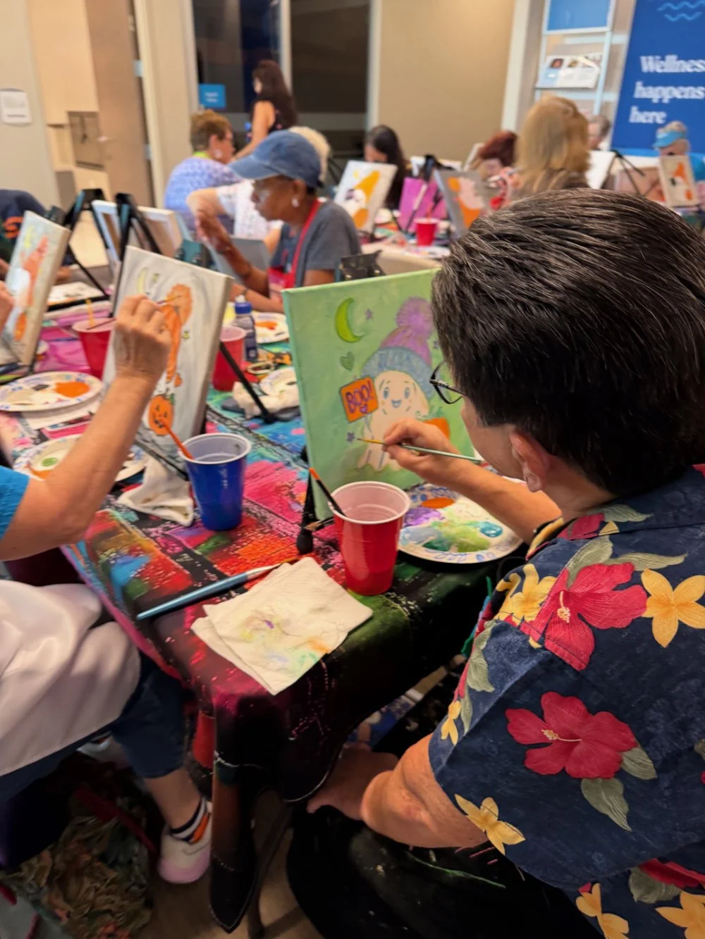 People participating in a group painting activity at a colorful table, creating artwork on canvases.