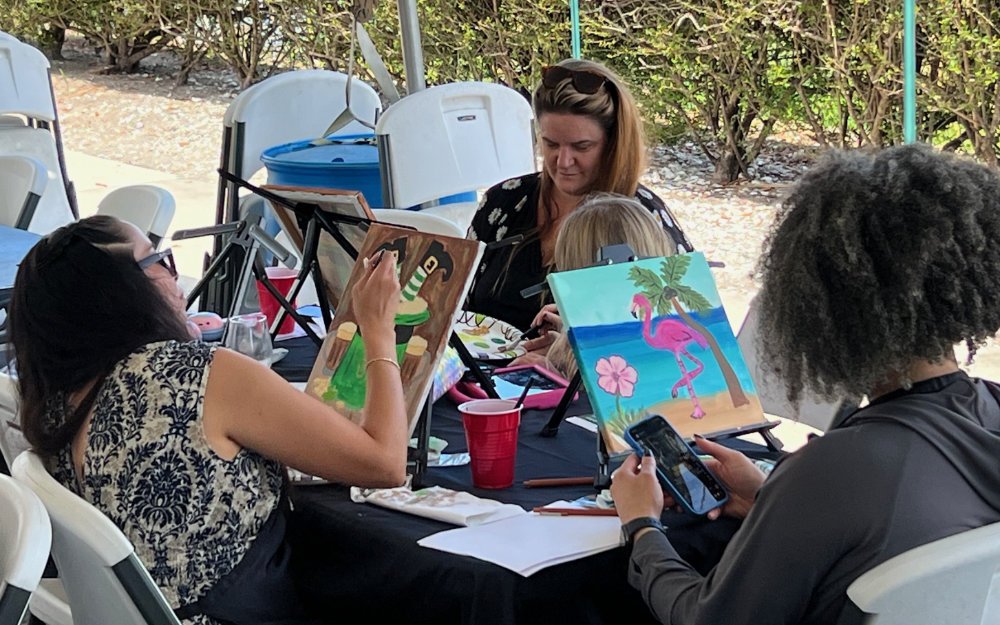 Four women participating in a painting class outdoors, creating tropical-themed artwork with a flamingo, palm trees, and beach scene.