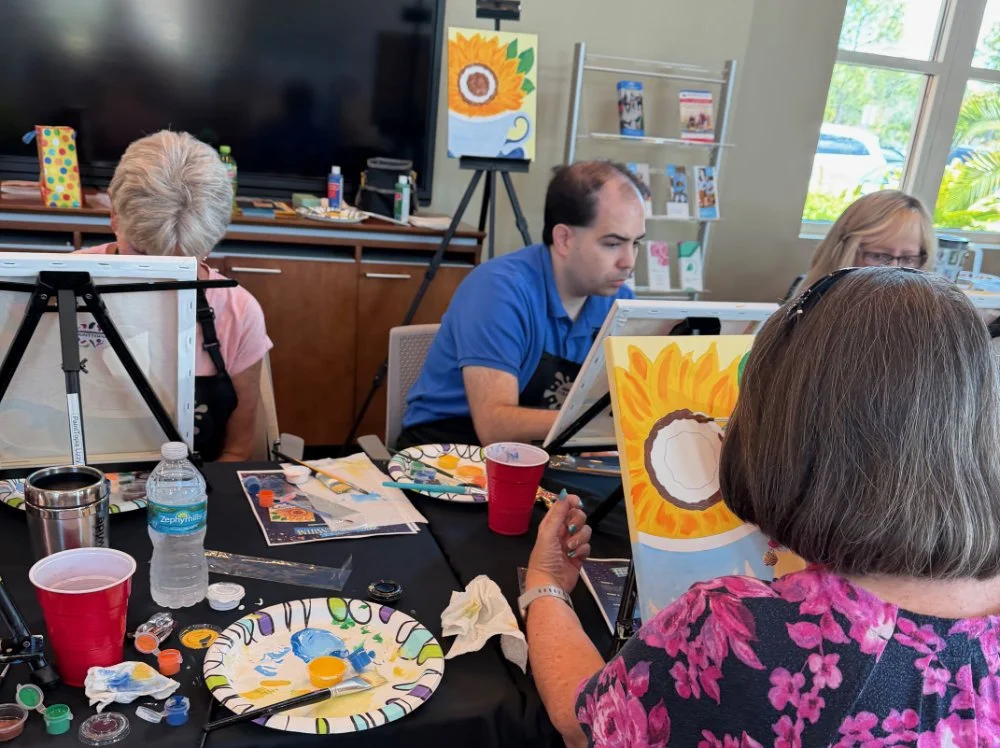 Adults and children painting sunflower pictures in an art class.