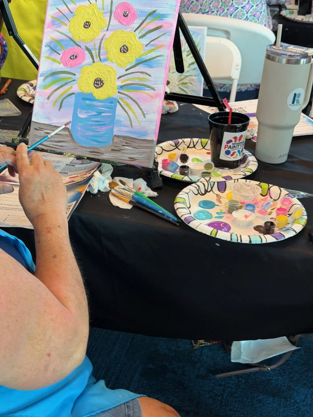 A person is painting a colorful still life of yellow and pink flowers in a blue vase on a canvas, surrounded by painting supplies, paints, brushes, and a painted paper plate with various colors on a black table.