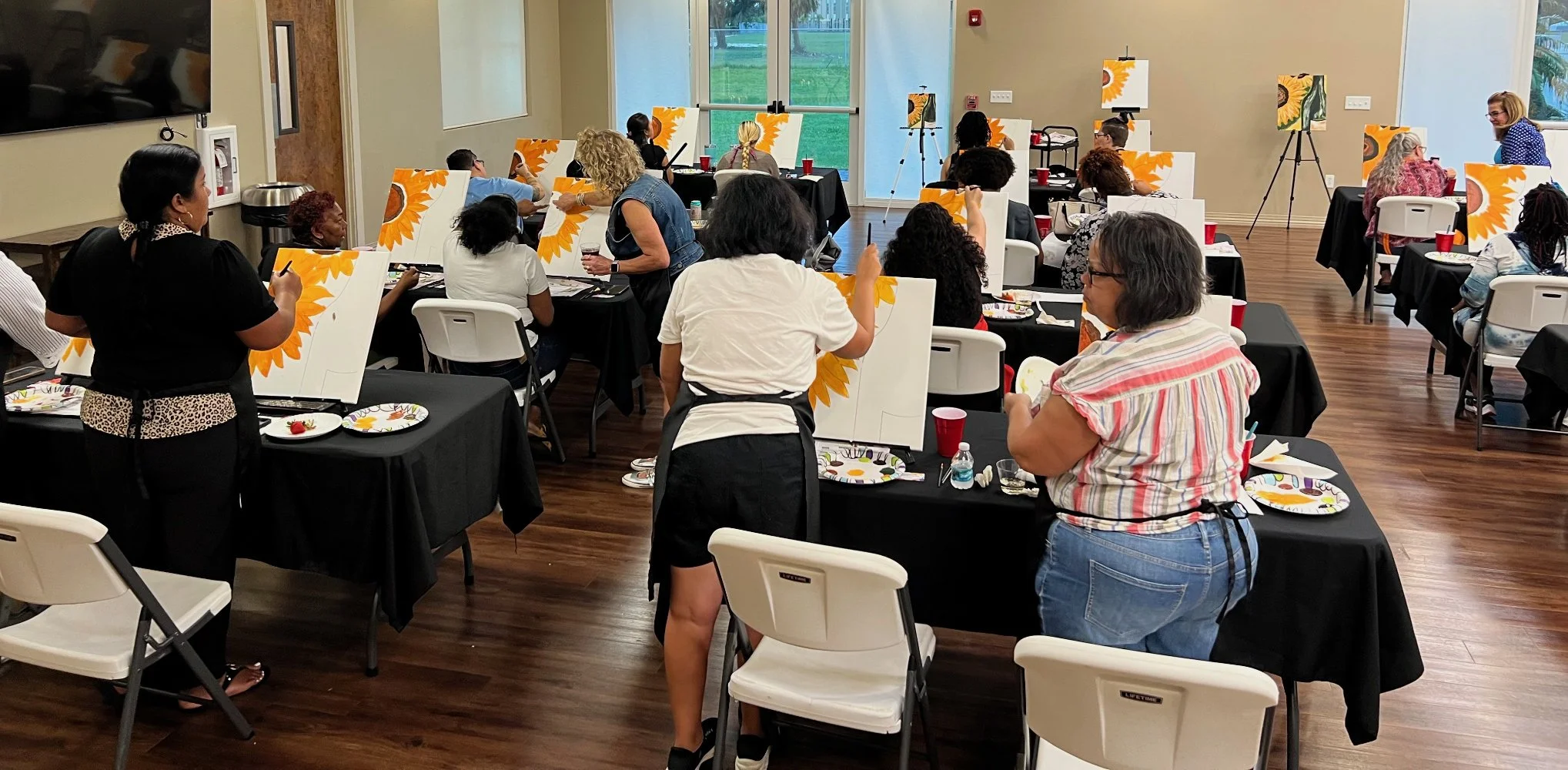 A group of people participating in an acrylic painting class in a well-lit room with large windows. The instructor is guiding a woman at the front, while others are working on their paintings at tables covered with black tablecloths. Each student has