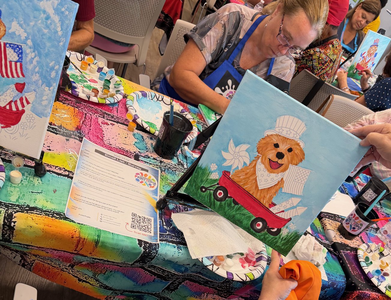 People participating in a paint-and-sip event, painting a colorful picture of a cute dog with a bandana and a hat on a canvas, with various painting supplies on the table.