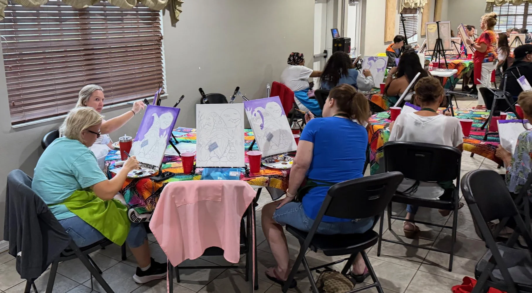 People participating in a group painting class, sitting at tables with art supplies, creating colorful animal-themed paintings in a well-lit room with large windows and wooden blinds.