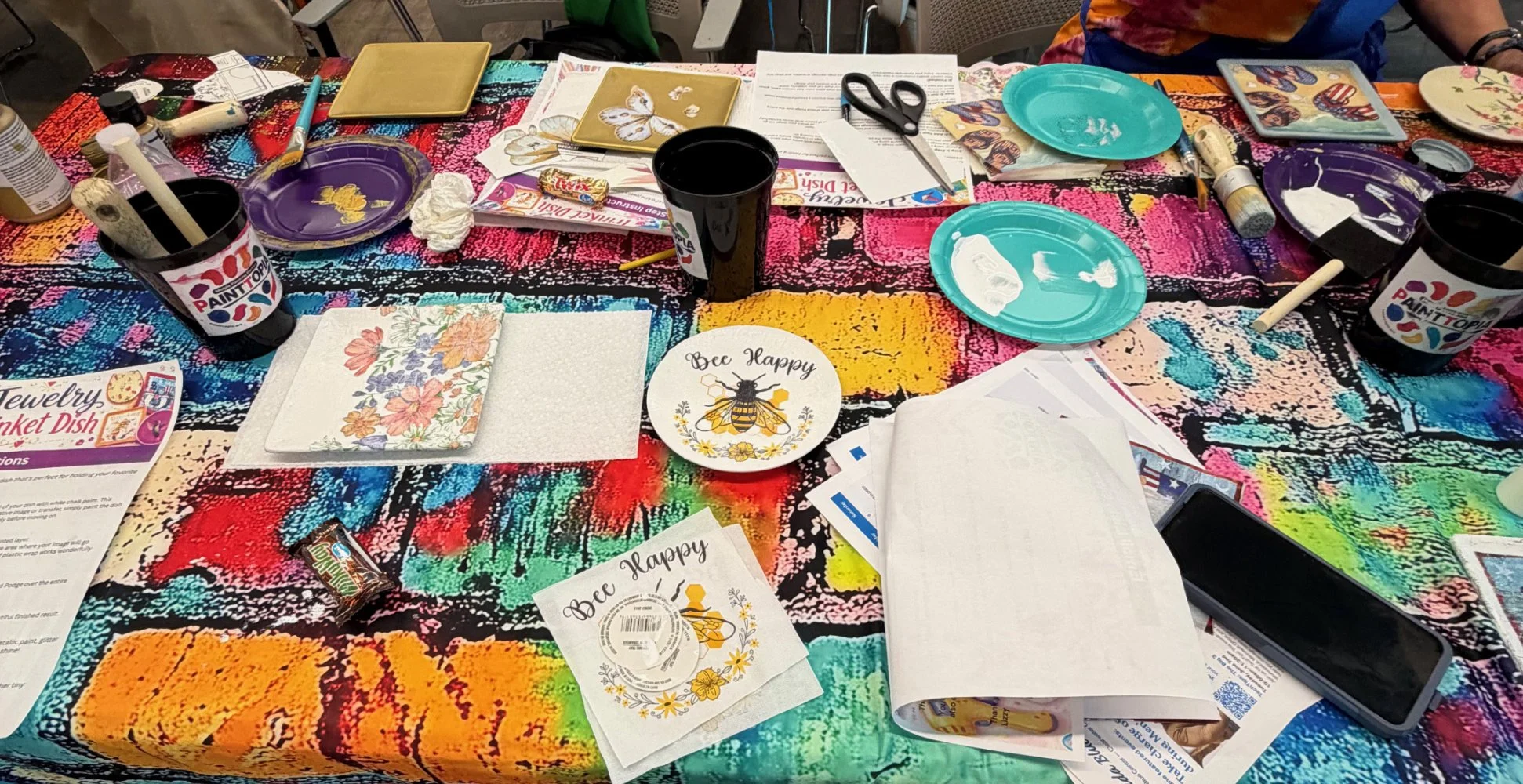 A colorful tablecloth covers a table filled with art supplies, paper notes, paints, brushes, plates, and scissors, suggesting an arts and crafts activity setup.